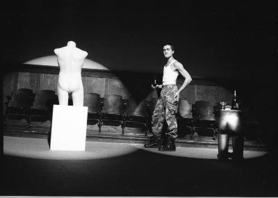 One spotlight gleams on the torso of a mannequin, positioned on a white plinth, while another lights up the bottom half of a mannequin functioning as a table, holding a bottle and glass. A performer in camouflage pants stands where the lights meet, with one hand on their hip and the other on the bristle end of a brush.
