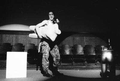  A performer in camo pants sings into a hairbrush while holding the mannequin by the hip in front of their body. They stand between a plinth and the bottom half of another mannequin.
