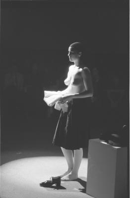 Jeanette Reinhardt stands under a spotlight in the Grand Luxe Hall in a state of undress. With her bare chest exposed, she faces the audience in profile while holding a white folded shirt. She wears stockings, a black pleated skirt, and a tight black head covering. One of her Mary Jane shoes is visible on the carpet next to her. 