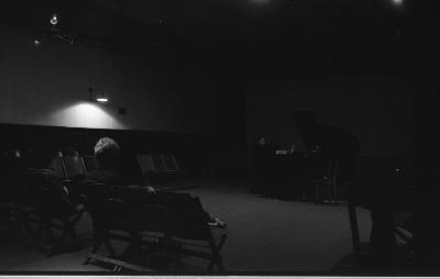 Two audience members sit in a low-light room, with a perimeter of otherwise empty chairs. They direct their attention towards a musician playing in the darkness.
