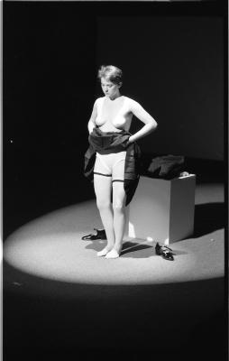 Jeanette Reinhardt stands topless under a spotlight and gazes at the carpeted floor. She lifts her skirt to reveal her undergarments and stockings. Her patent black mary jane shoes are on the floor next to her, and the rest of her clothes are stacked on a plinth behind her.