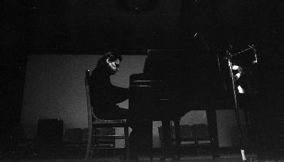 A darkened room is captured by a low-angle shot of a towering musician dressed in black bleeding into the darkness of an even larger grand piano. Mimicking the curvature of the wicker chair in which they sit, the artist bends into the piano.
