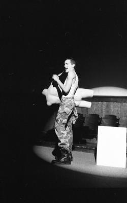 A performer in camo pants sings into a hairbrush while tucking the torso of a mannequin under their arm and behind their body. They perform in high contrast lighting, where everything lit is bright and everything outside the beams is deep black.
