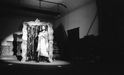 A performer stands in front of a sculptural portal made with a wavy frame and shimmering streamers which reach the floor. The performer crosses their arms, wears a flowy dress with a deep cut neck and black boots with a heel.
