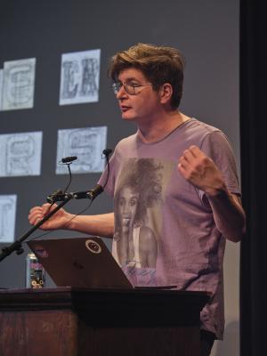 Wearing a purple Whitney Houston t-shirt, Dallas Hunt stands behind a lectern in the Grand Luxe Hall, gesturing idiosyncratically while speaking into a microphone. A can of craft beer sits next to his open laptop on the lectern. A projected image by Christian Vistian is visible in the background.