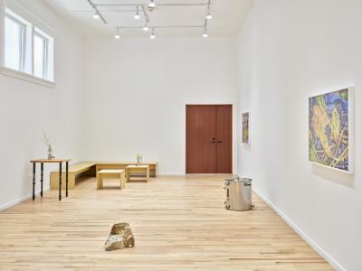 An installation view of a white-walled art gallery. There are two framed photographs hung along the right wall. On the gallery floor, there is an altered brewing unitank and a polka dot agate sculpture. In the far corner, there are a series of wooden benches and tables, some of which have floral arrangements sitting on them. Natural light shines in through two windows on the right wall, and the wooden gallery doors are closed.
