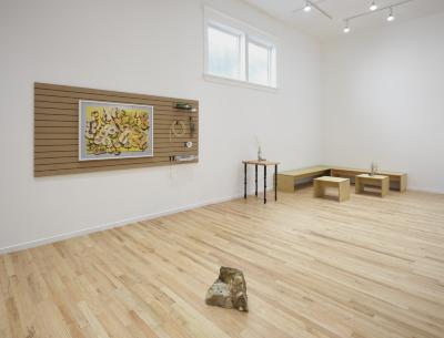 An installation view of a white-walled art gallery. Various brewing related objects along with a framed photograph of sliced apples on a yellow cutting board are hung on a slatwall panel on the left wall. In the far corner, there are a series of wooden benches and tables, some of which have floral arrangements sitting on them. A polka dot agate sculpture sits in the centre of the room. Natural light shines through two windows.