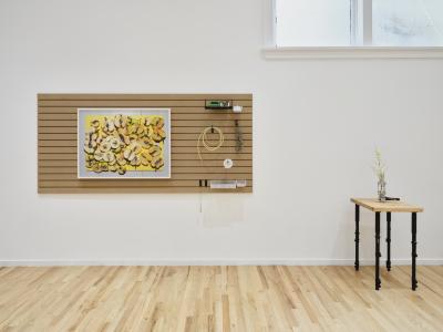 Various brewing related objects along with a framed photograph of sliced apples on a yellow cutting board are hung on a slatwall panel on a white gallery wall. To its right, a small floral arrangement in a vase made of a sight glass and metal brewing valve sits on a table made of a cutting board and adjustable monopod legs.
