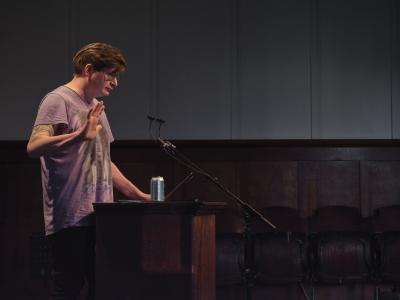 Dallas Hunt stands behind a lectern affixed with a microphone in the Grand Luxe Hall. He gazes towards the audience while holding up his right palm. A can of craft beer is positioned on the lectern next to his open laptop.