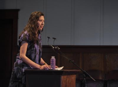 Leanne Dunic stands behind a lectern affixed with a microphone in the Grand Luxe Hall. She looks out towards the audience while reading from a set of printouts. Her purple water bottle sits open on the surface of the lectern. 