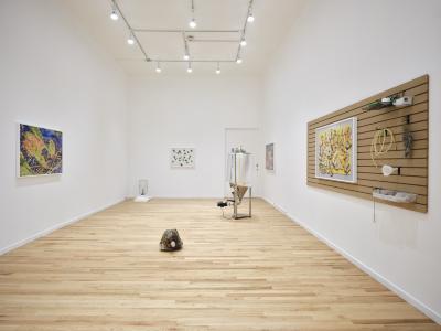 A wide angle installation view of a white-walled art gallery. Several photographs hang on the walls, one of which is on a slatwall panel alongside various brewing related equipment.  Brew-related sculptures are interspersed on the floor.