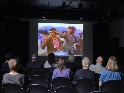 Margaret Dragu and Kate Craig’s 1979 video work Backup is projected on a screen in the Grand Luxe Hall. The still shows Craig and Dragu standing on the roof of Western Front, pointing beyond the camera’s frame. The first four rows of audience members are visible from behind.