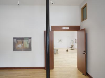 A framed photograph of grain in a mash tun hanging on a white wall outside of a gallery with its doors open. Inside the gallery, there are several photographs and brew-related sculptures.