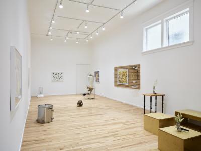 A wide angle installation view of a white-walled art gallery. Several photographs hang on the walls, with wooden tables, benches and brew-related sculptures interspersed on the floor. Track lights line the ceiling, as natural light comes through two large windows.