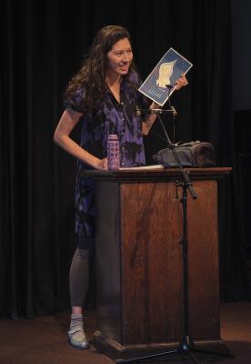 Leanne Dunic stands behind a lectern affixed with a microphone in the Grand Luxe Hall. She gazes towards the audience while loosely holding a stack of paper in her left hand. The pages rest against the surface of the lectern next to a purple water bottle.