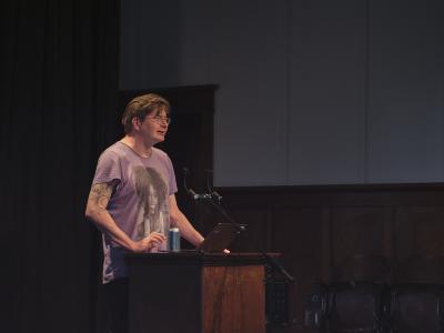 Wearing a purple Whitney Houston t-shirt, Dallas Hunt stands behind a lectern affixed with a microphone in the Grand Luxe Hall. A can of craft beer is positioned on the lectern next to his open laptop.