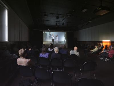 Margaret Dragu’s 2023 video work Tick and Talk of Common Time is projected on a screen in the Grand Luxe Hall. The image on the screen shows Dragu and Justine A. Chambers dancing in a large studio with cement floors. The audience in the room is visible from behind. Light refracts through a nearby window and leaves a diffused pattern on the far wall.
