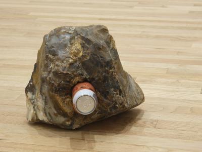 A can of beer with an orange label fits snuggly into a hole in a piece of polka dot agate placed on a wooden floor.