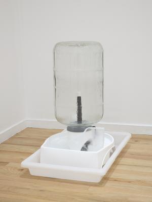 A glass carboy receives a blast of water from a keg washer on which it is placed. A piece of selenite sits in the keg washer’s basin. The entirety of the sculpture sits in a white, shallow plastic tray placed in a corner of an art gallery.
