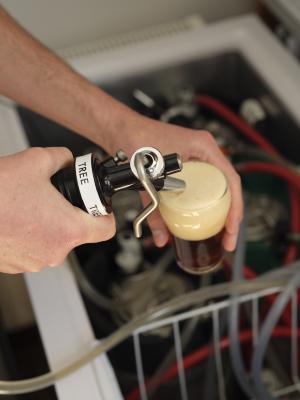 A close up image of a pair of hands pouring beer from a tap into a glass. The left hand holds the glass with beer foaming to its rim, while the right holds a trigger-style tap labeled with the word “TREE”. In the background, out of focus, are several kegs and lines in an open chest freezer.