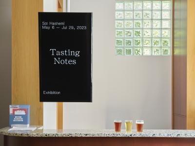 A well-lit reception area with wood panels, white walls and glass block windows. A vertical monitor suspended over a terrazzo countertop reads “Sol Hashemi May 6 - Jul 29, 2023 Tasting Notes Exhibition”. A blue suggestion box and three small glasses of brews, two amber and one yellow in colour, sit on the countertop.