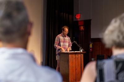 D.M. Bradford stands behind a lectern affixed with a microphone in the Grand Luxe Hall. They gaze down towards their reading material. The image frames D.M. between the backs of two audience members’ heads.