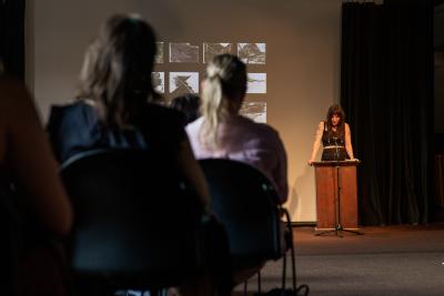 Danielle LaFrance stands behind a lectern affixed with a microphone while reading in the Grand Luxe Hall. With their elbows locked, they grip the edges of the lectern as they cast their gaze downwards. Danielle is positioned in front of a projected image by Christian Vistan that presents twenty of black-and-white abstract paintings arranged in a grid of five columns and four rows. Seated in rows, the audience is visible from behind.