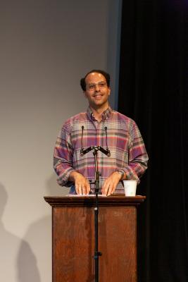 D.M. Bradford stands behind a lectern affixed with a microphone in the Grand Lux Hall. Their fingertips rest on top of their reading material as they smile towards the audience.