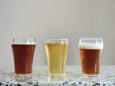 Three evenly spaced glasses on a terrazzo countertop. Each is filled within a centimeter of the top with brew. The middle brew is of a cider light yellow colour, and is flanked by two amber coloured beers.