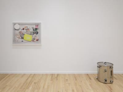 A framed photograph of a variety of rocks, gemstones and brightly-colored plastics placed in a white bussing tray hangs on a white gallery wall. Adjacent to it, on the floor to the right, is a stainless steel kettle with custom fittings.