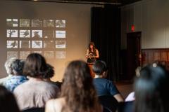Standing behind a lectern affixed with a microphone in the Grand Luxe Hall, Danielle LaFrance reads from their book #postdildo. They are positioned in front of a projected image by Christian Vistan that presents twenty of black-and-white abstract paintings arranged in a grid of five columns and four rows. The first five rows of audience members are visible from behind.