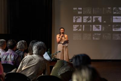 Deanna Fong stands facing the audience in the Grand Luxe Hall. Smiling, she holds a cordless microphone up to her mouth with her right hand, while her left hand holds her speaking notes. She is positioned in front of a projected image by Christian Vistan that presents a number of black-and-white abstract paintings arranged in a grid. Seated in rows, audience members can be seen from the back.