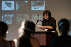 Jane Shi stands at a lectern affixed with a microphone in the Grand Luxe Hall. She reads from a piece of paper held with both hands along its bottom edges. Jane is positioned in front of a projected image by Christian Vistan that presents a number of black-and-white abstract paintings arranged in a grid. The first row of audience members are seen from behind.