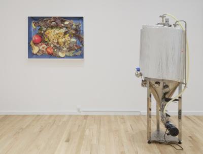 A framed photograph of apples and brown leaves in a blue plastic bin hangs on a white gallery wall. Adjacent to it, on the right, is a brew-sculpture of an insulated unitank, complete with a pump, hose, and custom fittings.