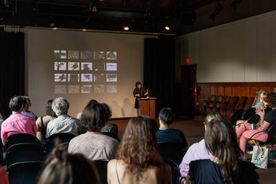 Jane Shi reads from a lectern affixed with a microphone in the Grand Luxe Hall. She is positioned in front of a projected image by Christian Vistan that presents twenty of black-and-white abstract paintings arranged in a grid of five columns and four rows. The first five rows of audience members are visible from behind.