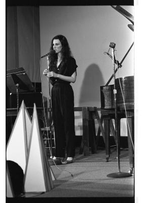 A long-shot of a woman with long hair dressed in a jumpsuit. She performs on stage as she speaks into a microphone. Behind her are several pieces of music equipment and a grand piano.