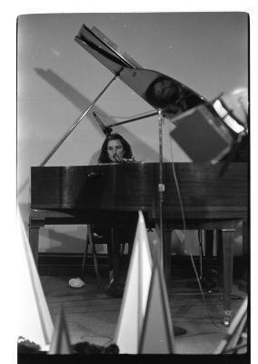 A front shot of a woman singing as she plays the grand piano. The foreground depicts blurred cone-like props and part of a stage light.