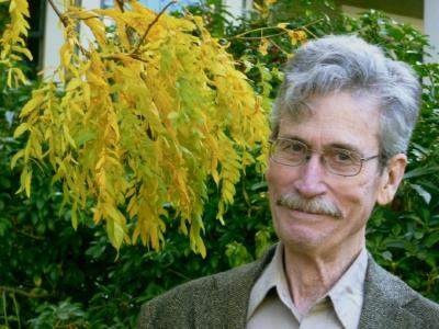 Patrick Ready stands next to a tree with yellow leaves. He wears wire framed glasses and a beige collared shirt under a grey tweed blazer. 