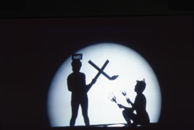 Two figures appear as shadows at the centre of a spotlight. One person stands holding a burning cross, while a devil figure with their tongue sticking out kneels in front while holding two large forks.