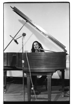 A front shot of a woman singing as she plays the grand piano. The foreground depicts a blurred microphone on a stand.