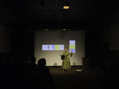 Tawhida Tanya Evanson reads from a black portfolio in her right hand, while her left hand energetically gestures upwards. She is barefoot in the Grand Luxe Hall, and wears a green and gold dress. A drum is positioned on the floor, and a projected image of paintings by Christian Vistan can be seen behind her.