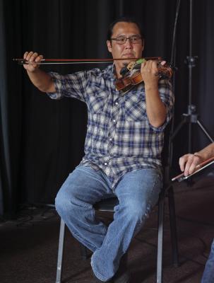 Wesley Hardisty plays violin while seated. He wears square glasses, a blue plaid shirt, and blue jeans.