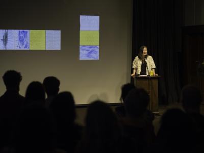 Isabella Wang addresses the audience in the Grand Luxe Hall from behind a lectern. Behind her is a projection of scanned paintings by Christian Vistan. The first few rows of the audience are visible from behind.