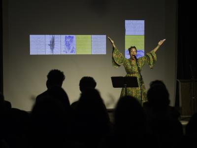 Tawhida Tanya Evanson’s arms are positioned in an energetic V-shape as she vocalizes into a microphone in the Grand Luxe Hall. She wears a gold and green patterned dress, and stands behind a black music stand. A projected image of abstract paintings by Christian Vistan can be seen in the background. The first row of the audience can also be seen from behind.