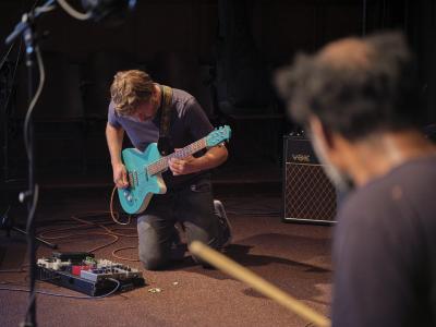 John Dieterich kneels on the carpet of the Grand Luxe Hall while playing a turquoise electric guitar. He gazes down at his collection of effects pedals, which are on the floor next to a pile of guitar picks. 