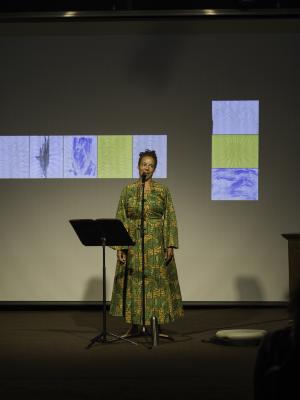 Tawhida Tanya Evanson smiles at the audience from behind a microphone in the Grand Luxe Hall. She wears a gold and green patterned dress and stands with her arms by her sides. A black music stand, metal thermos, and drum are positioned around her. A projected image of paintings by Christian Vistan can be seen in the background.