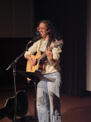 Sonnet L’Abbé plays acoustic guitar in the Grand Luxe Hall. They smile at the audience from behind their microphone and music stand. Sonnet wears orange glasses, a light green hoodie, and light wash blue jeans.