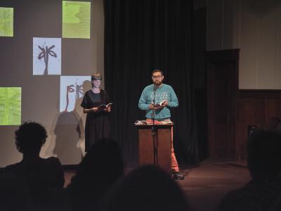 Klara du Plessis wears a black linen dress and gazes down at an open book she holds with both hands. To her right, Khashayar Mohammadi speaks into a microphone positioned in front of a lectern. They wear glasses, a blue sweatshirt, and orange pants, and hold an open book in their hands. They gaze towards the audience in the Grand Luxe Hall, some who are visible from behind. Klara and Khashayar Mohammadi stand in front of a projection with abstract, water-based paintings by Christian Vistan.