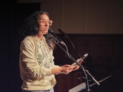 Sonnet L’Abbé speaks into a microphone affixed to a stand. They hold a tablet with both hands, and are wearing orange glasses, and a green hoodie with botanical illustrations and text reading Harm Reduction is Good…