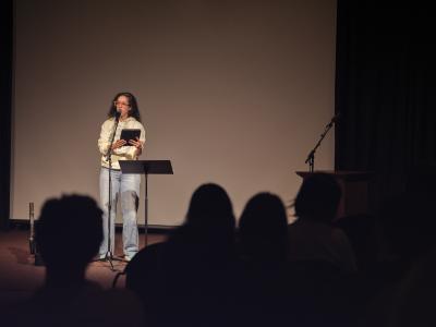 Sonnet L’Abbé speaks into a microphone on a stand in the Grand Luxe Hall. They hold a digital tablet in both hands, and wear orange glasses, a light green hoodie, and blue jeans. A black music stand is positioned next to them.